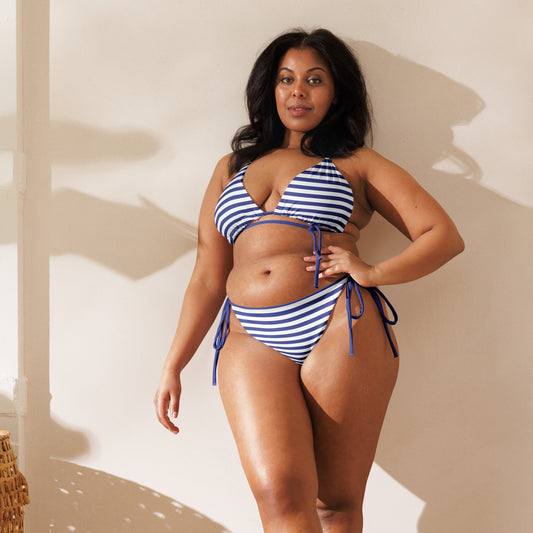 A curvy woman is wearing a navy and white striped string bikini, designed for a stylish day at the beach, paired with matching pet swimwear for eco-conscious beachgoers.